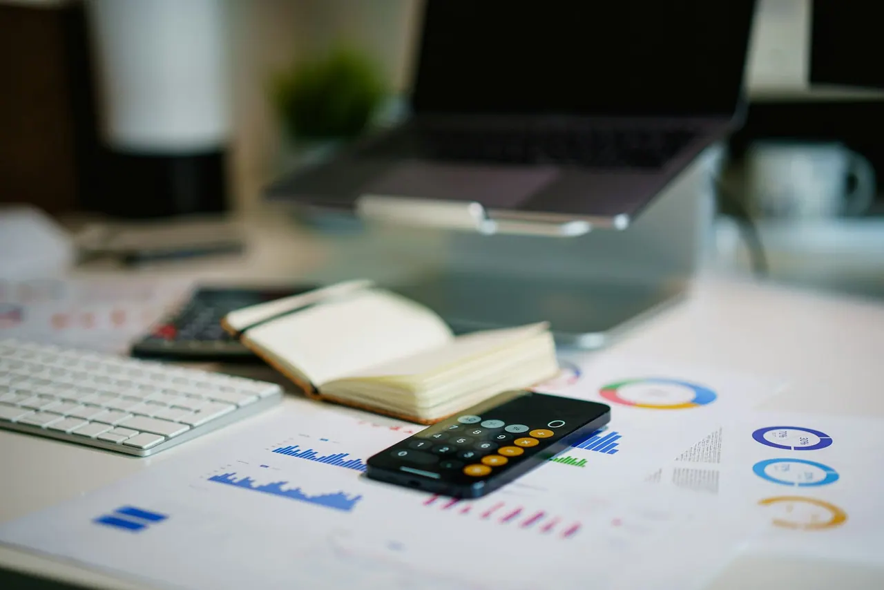 A desk with printed charts, a notebook and a calculator on a phone