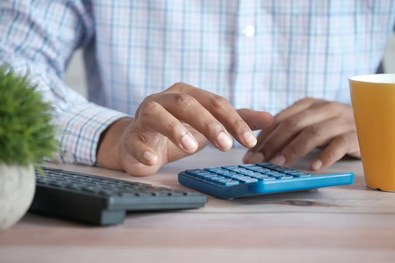 Hands working a calculator beside a keyboard and a yellow mug