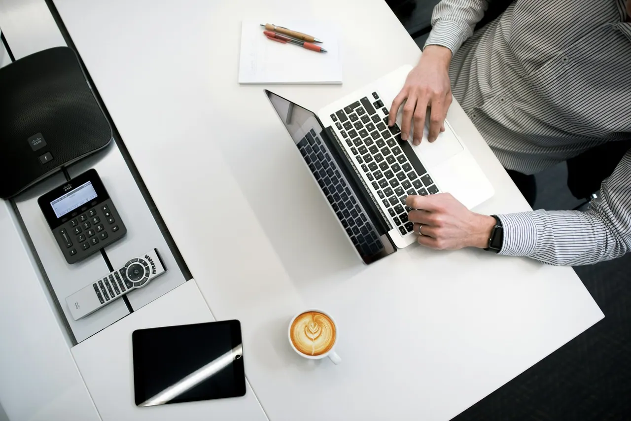 A tidy white desk from above with a laptop, a coffee and a card reader