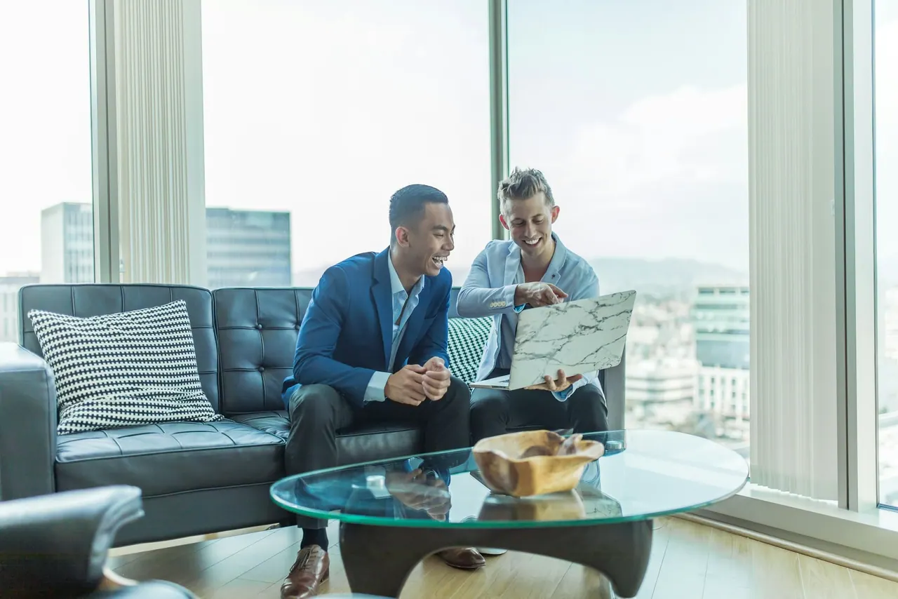 Two advisers laughing over a laptop on a leather sofa, by tall windows high above the city