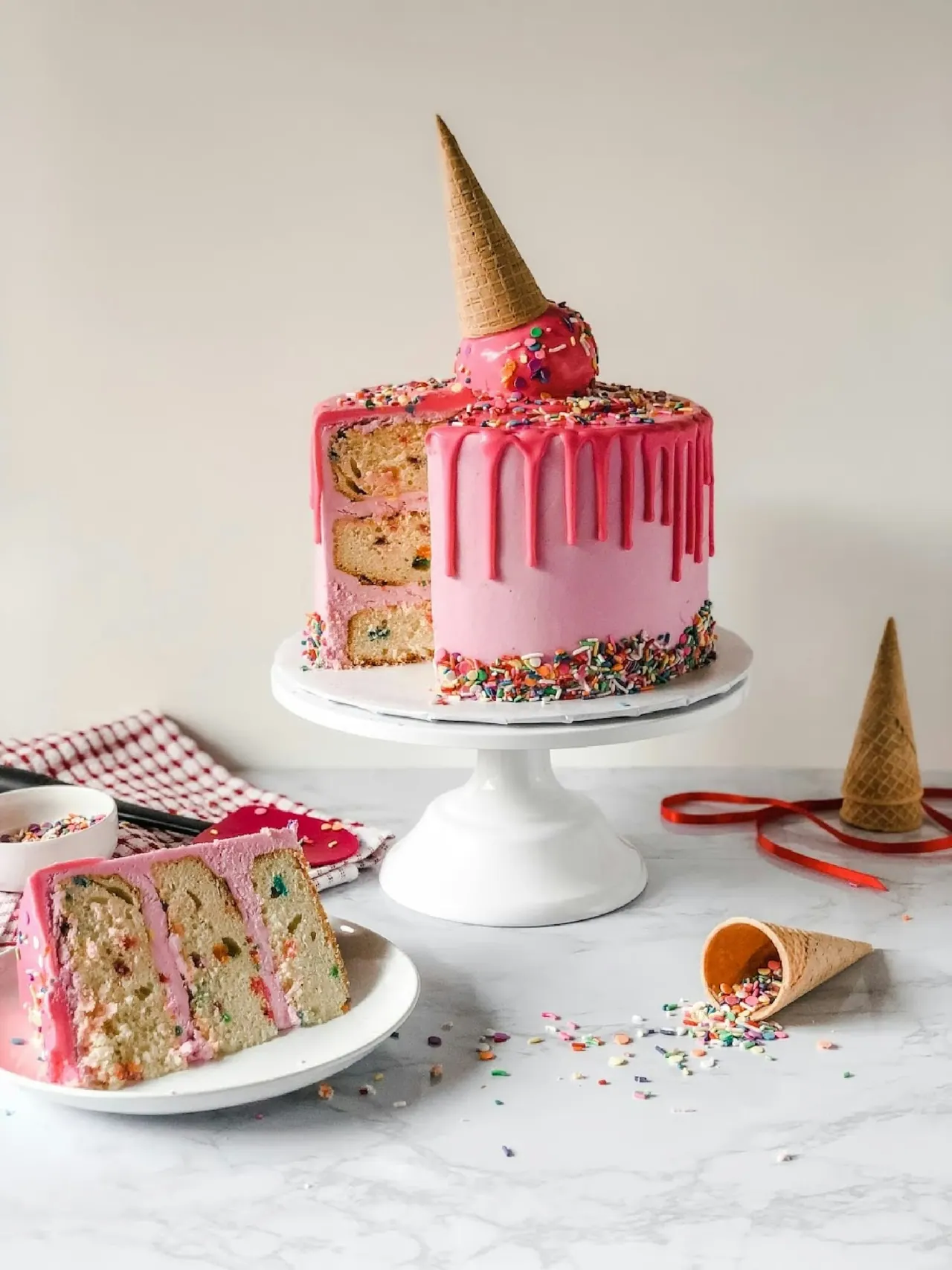 A pink drip cake with sprinkles and a cone on top, a slice beside it