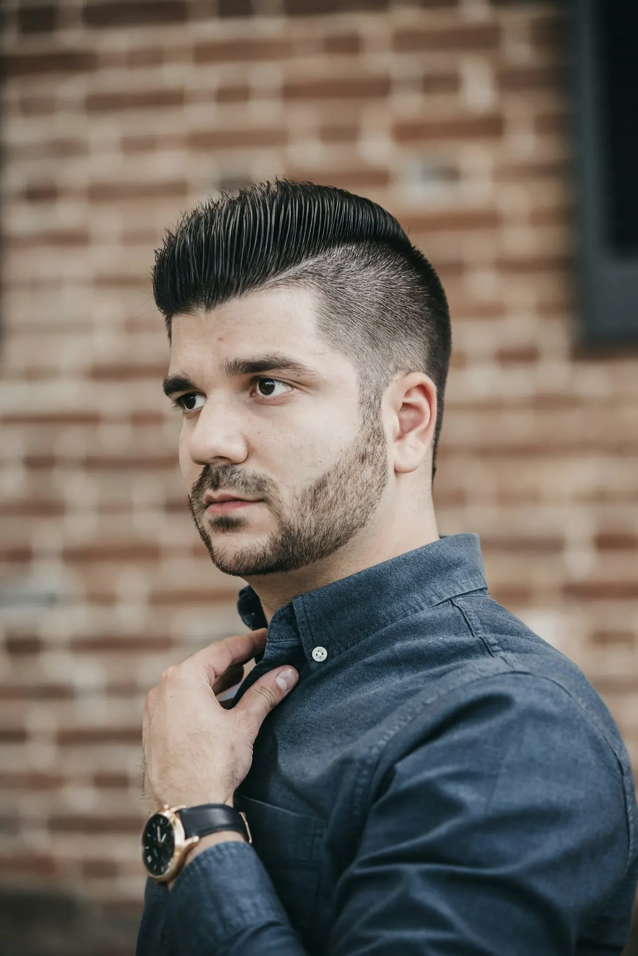 A man with slicked back hair and a sharp skin fade by a brick wall