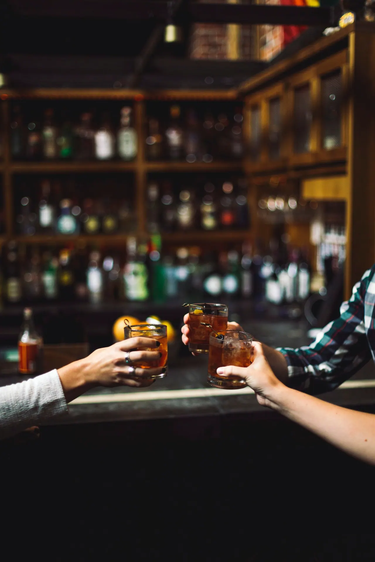 Two friends clinking glasses at a bar lined with bottles