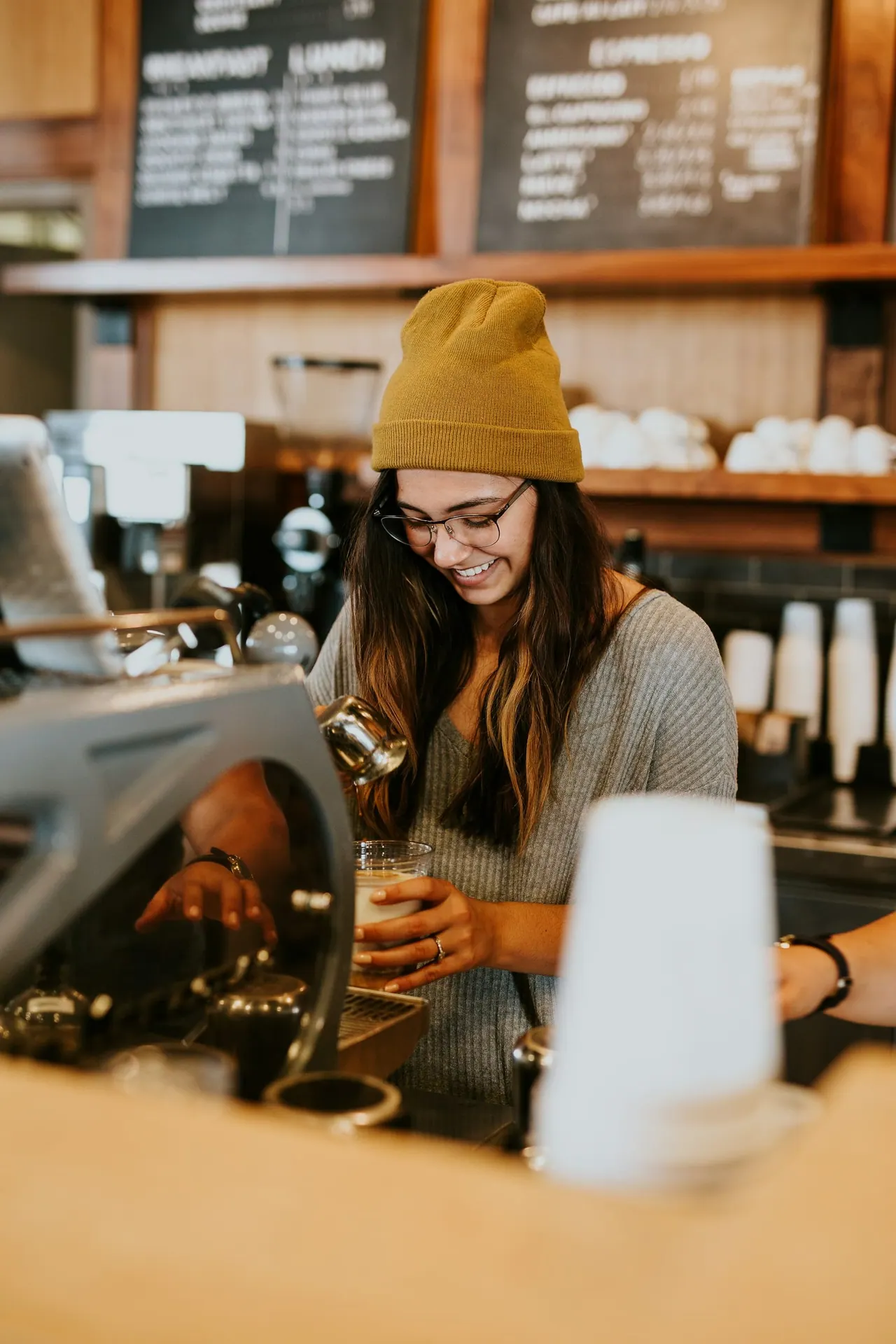 A smiling barista in a mustard beanie finishing a latte
