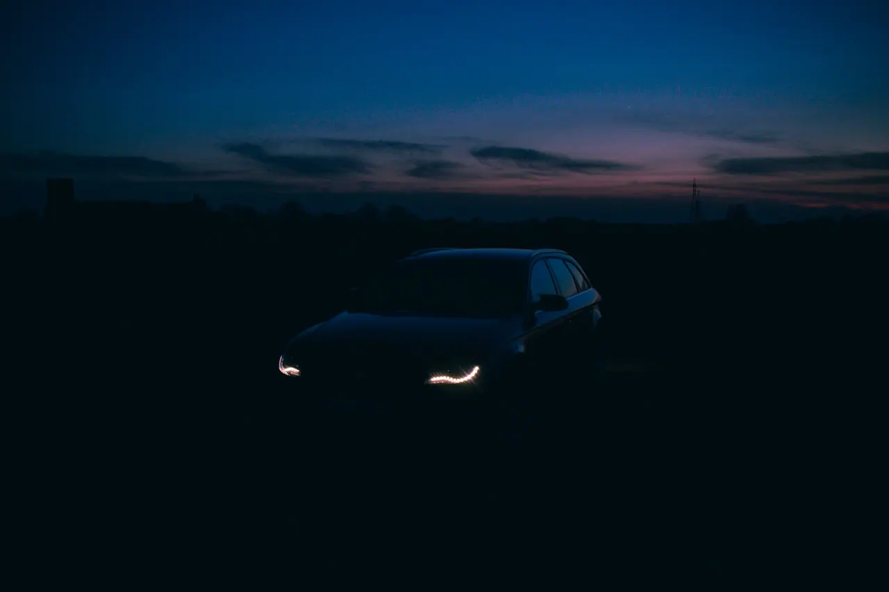 A dark sedan with its headlights on at dusk