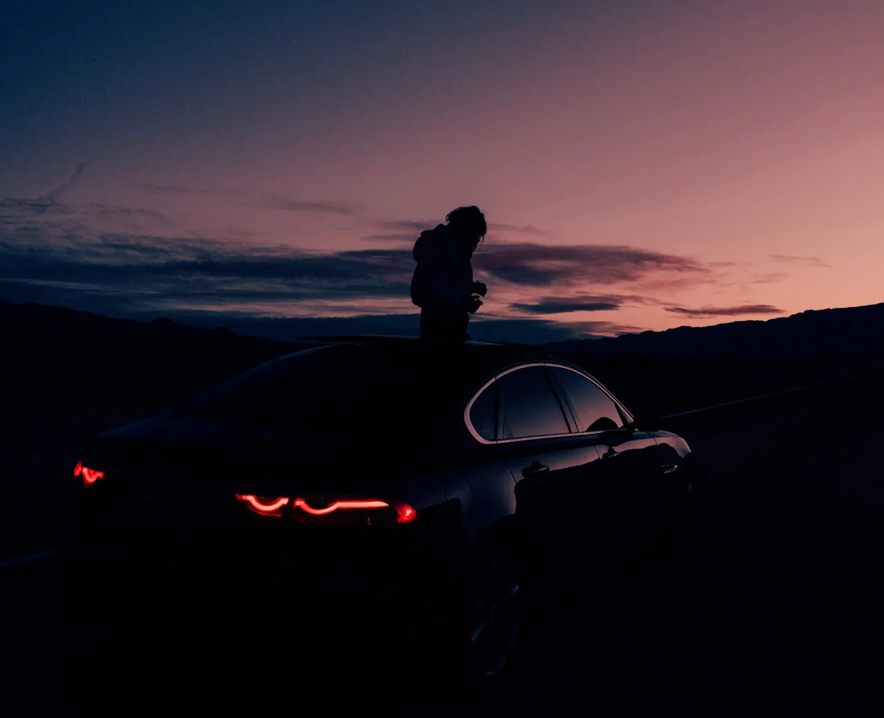 A person standing on the roof of a dark coupe at dusk