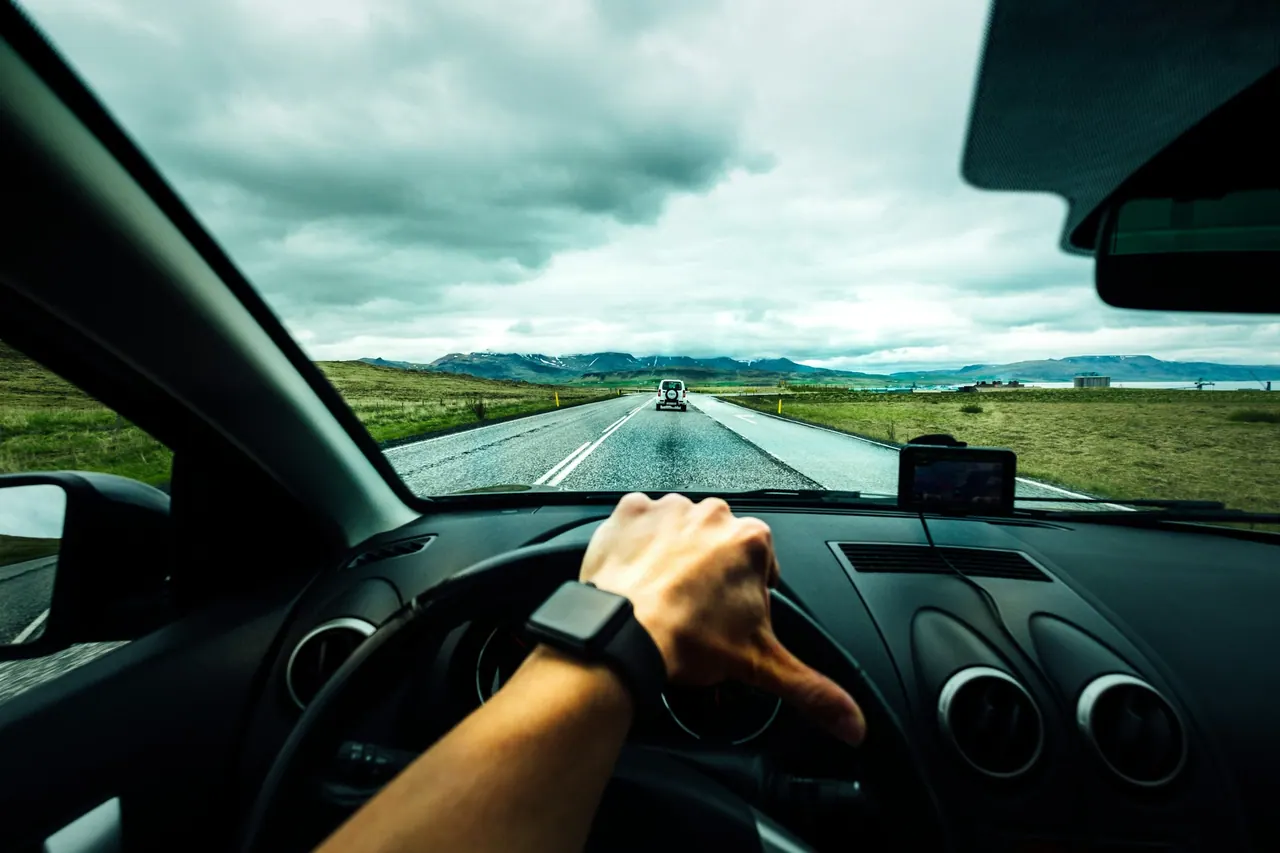 A hand on the steering wheel on an open road under a cloudy sky