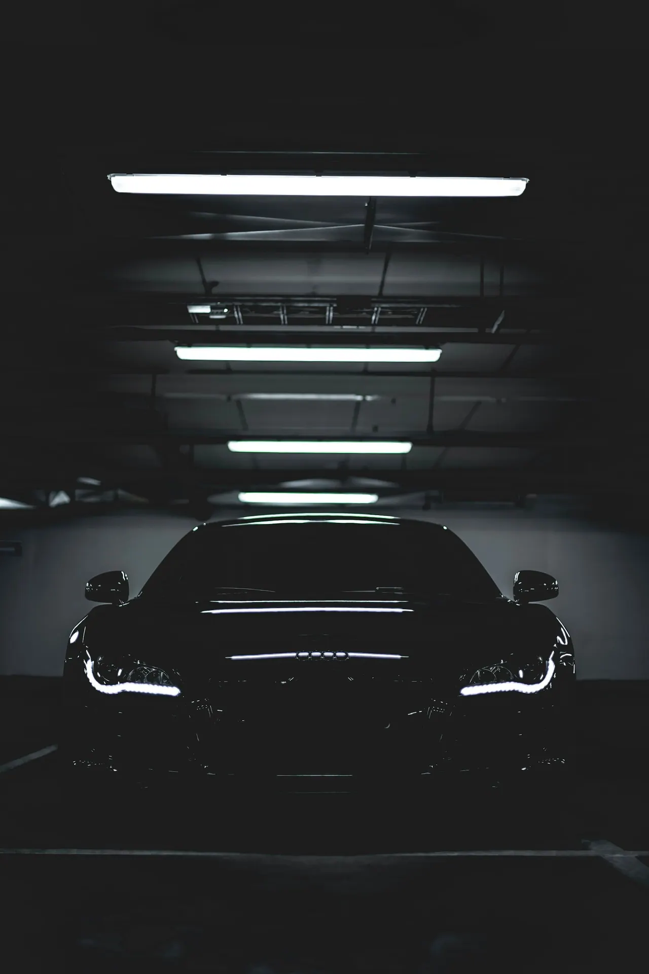 A dark coupe under strip lights in a garage, headlights on