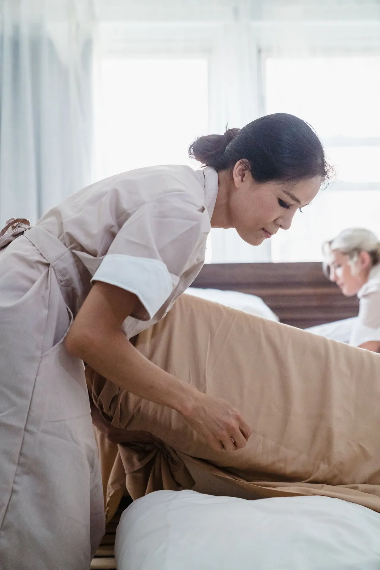 A cleaner smoothing a fresh sheet over a bed by a bright window