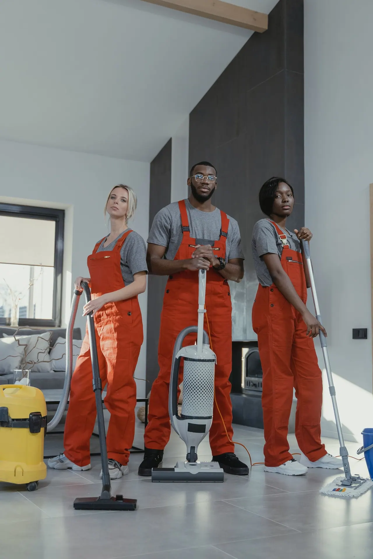 Three smiling cleaners in orange uniforms with a vacuum and mops in a bright home