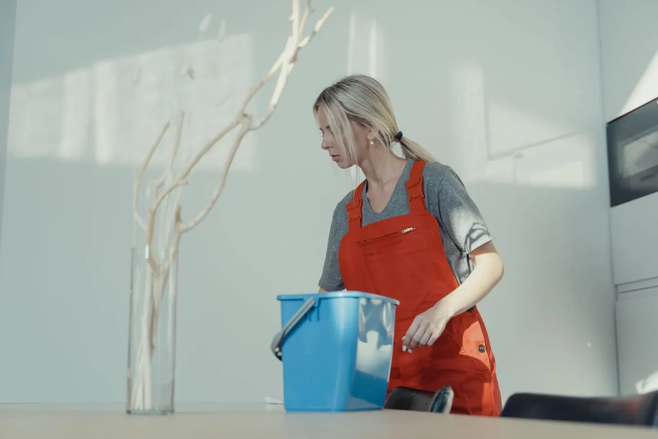 A cleaner in an orange apron carrying a blue bucket of supplies