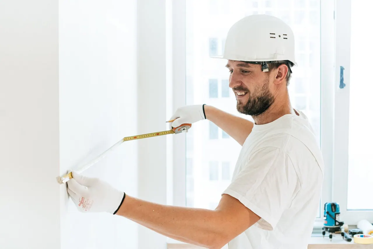 A smiling tiler in a hard hat measuring a wall with a tape