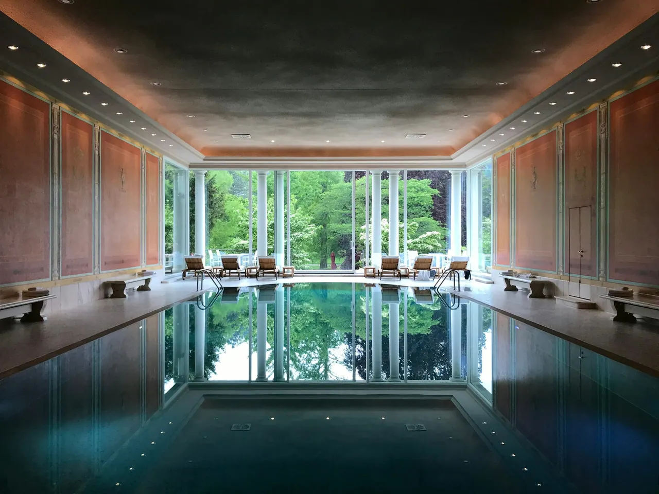 A grand indoor pool, still as glass, reflecting tall windows onto a garden