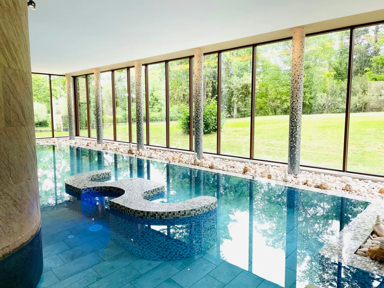 An indoor pool beside a wall of windows onto a green lawn