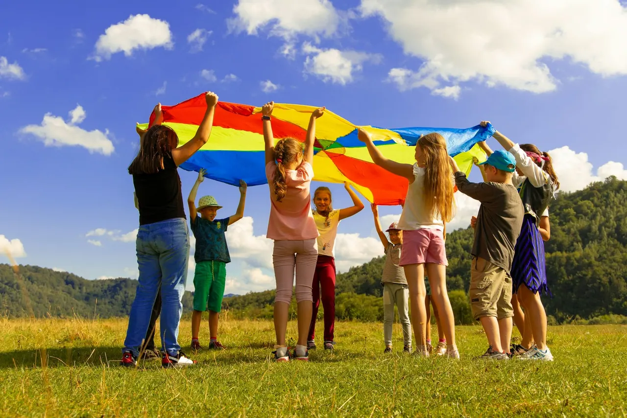 Children and teachers holding up a rainbow play parachute in a sunny field