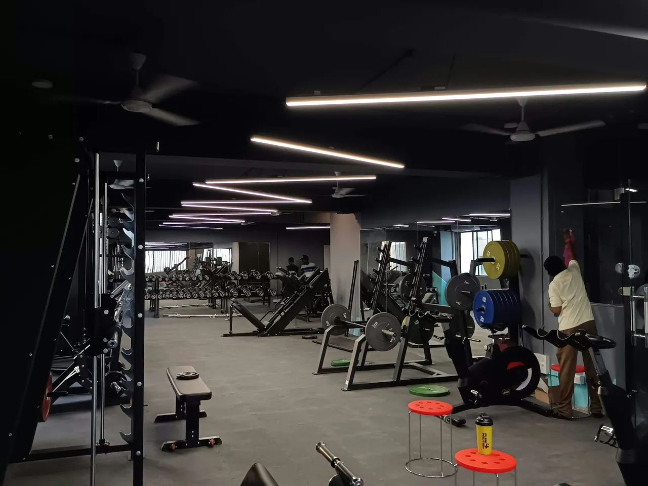 The gym floor under long strip lights, with racks and coloured plates