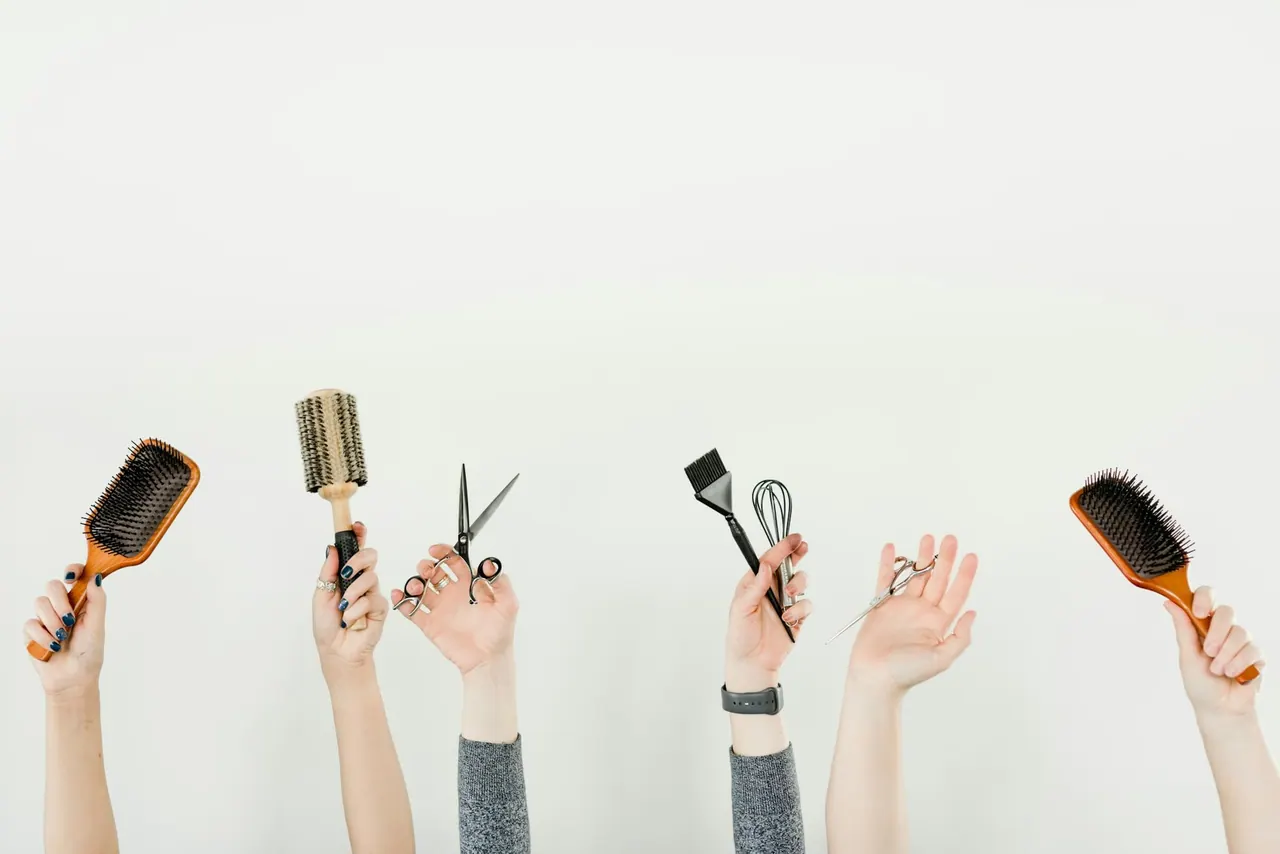 Hands holding up brushes, a comb and scissors against a white wall