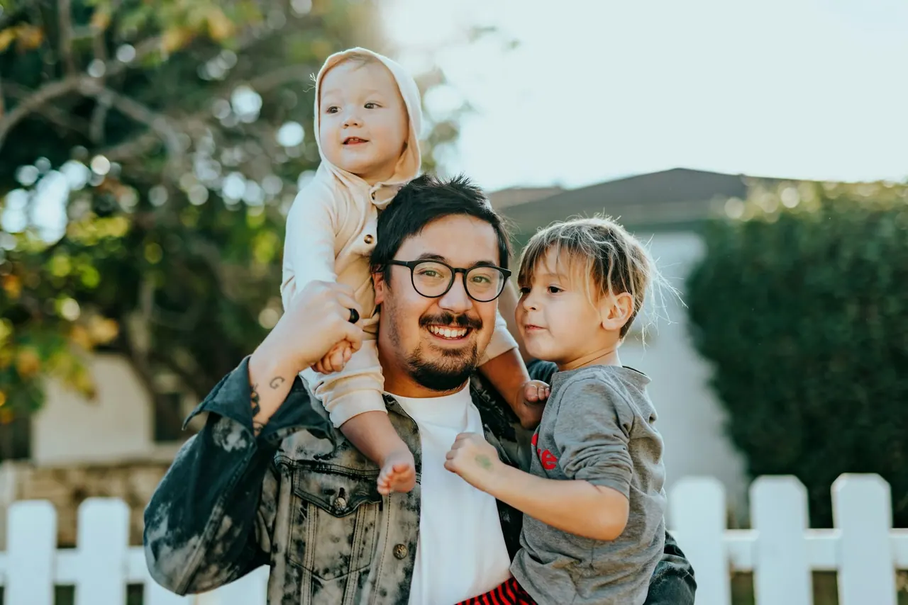 A smiling father carrying his two young children outside their home