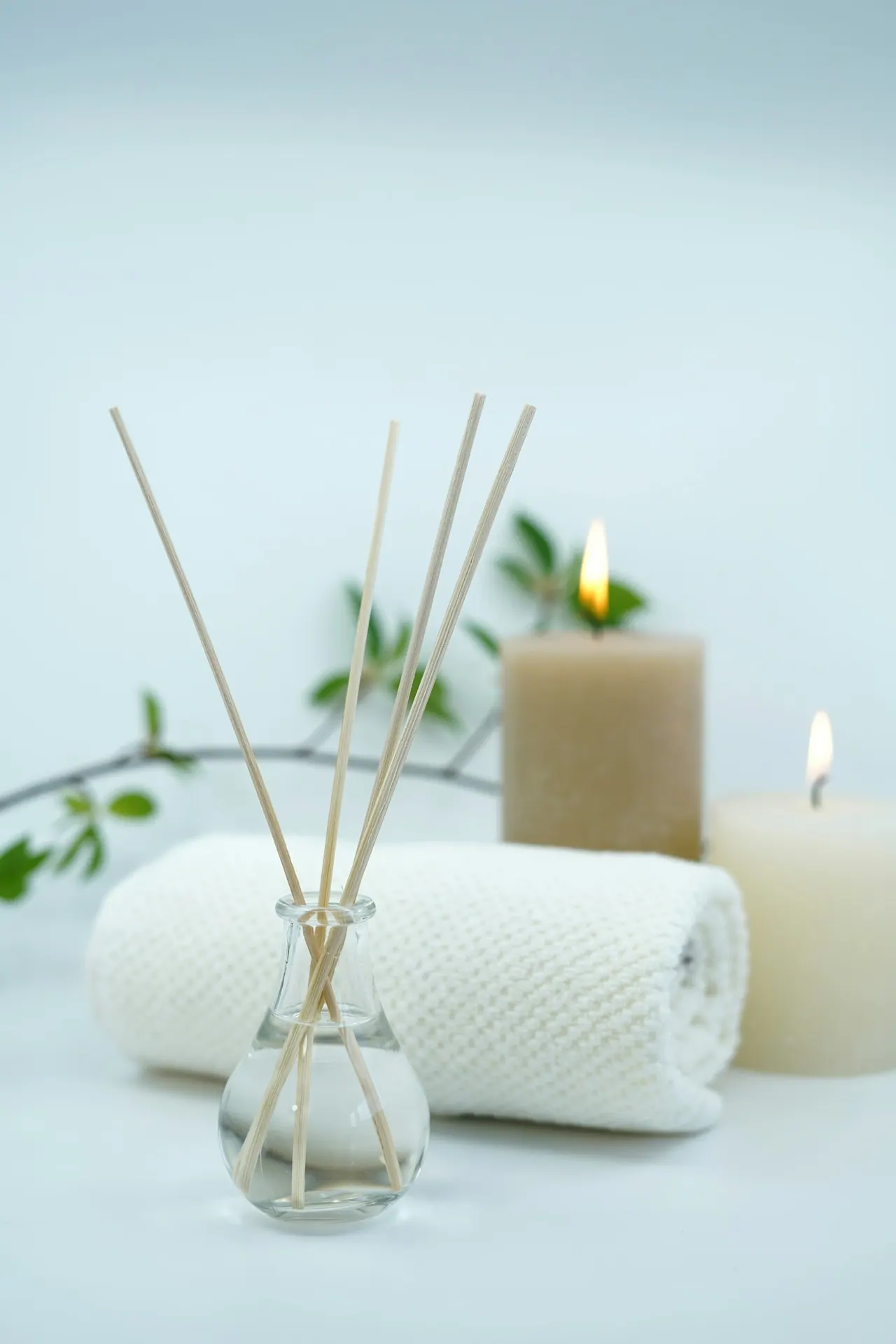A reed diffuser, a rolled white towel and two lit candles on pale blue