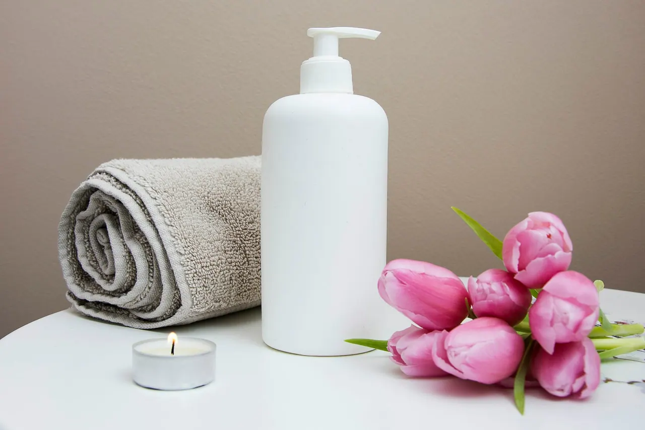 A white pump bottle of cream beside a rolled towel, a candle and pink tulips