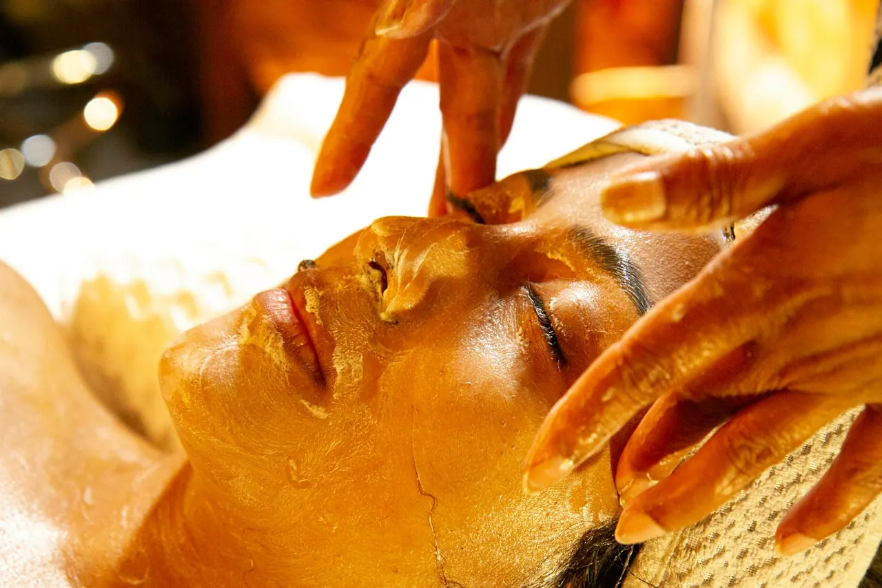 A therapist smoothing a golden mask over a client's face in warm light