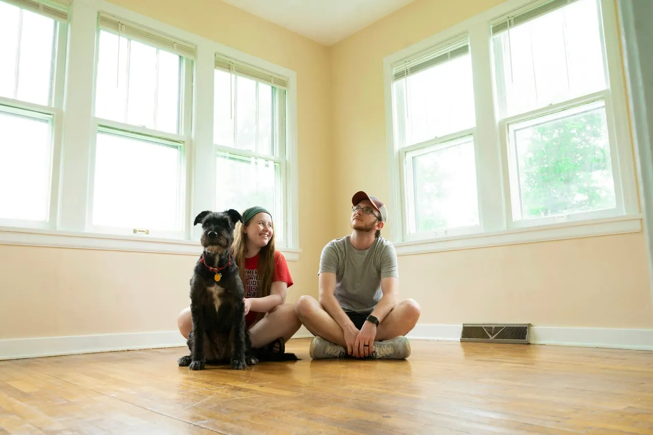A couple and their dog sitting on the wooden floor of an empty, sunny room