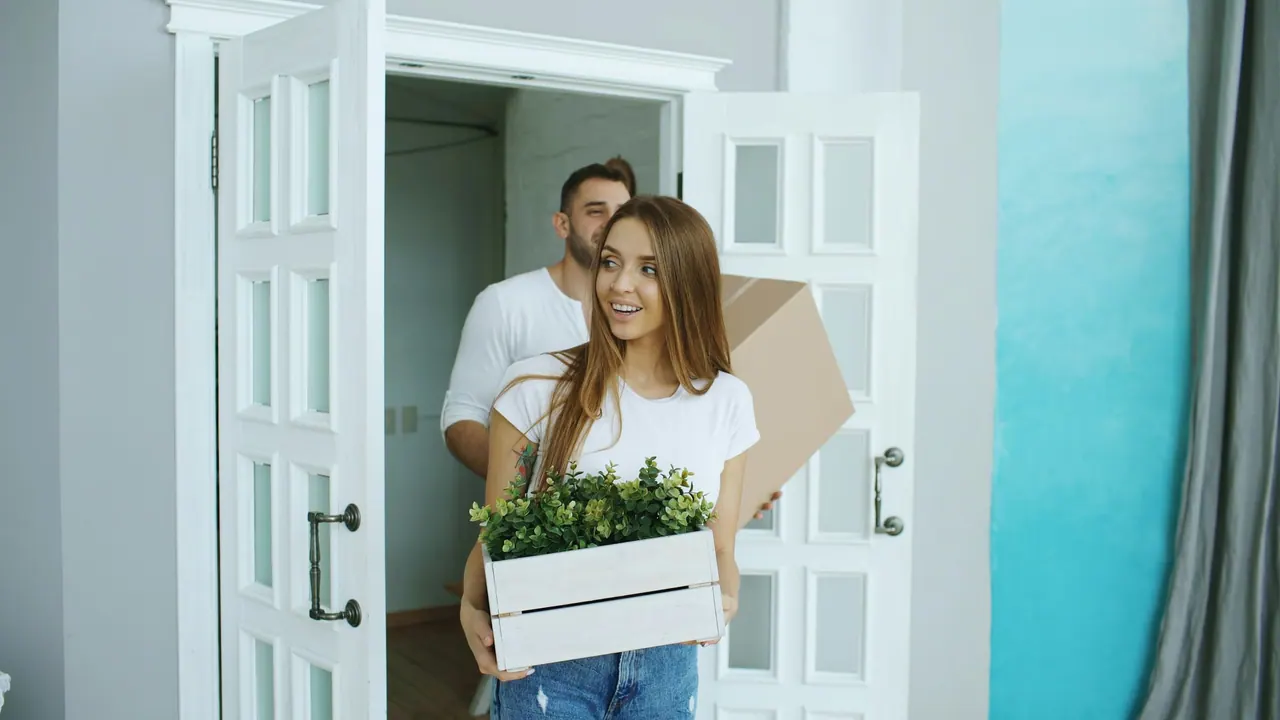 A smiling couple carrying a box and a crate of plants into their new home
