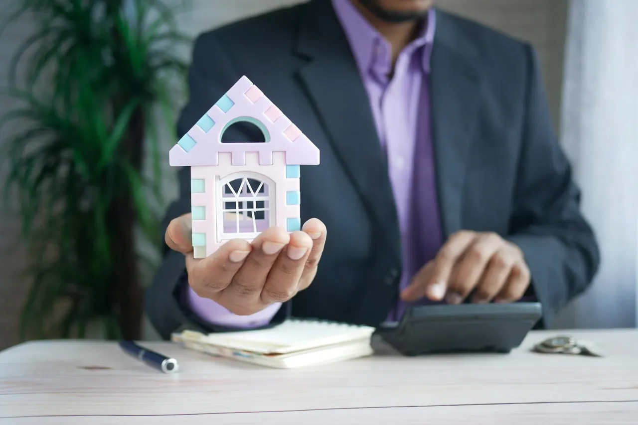 A broker at a desk holding a small model house beside a calculator
