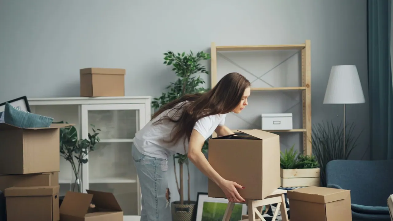 A woman lifting a cardboard box in a bright living room full of packed boxes