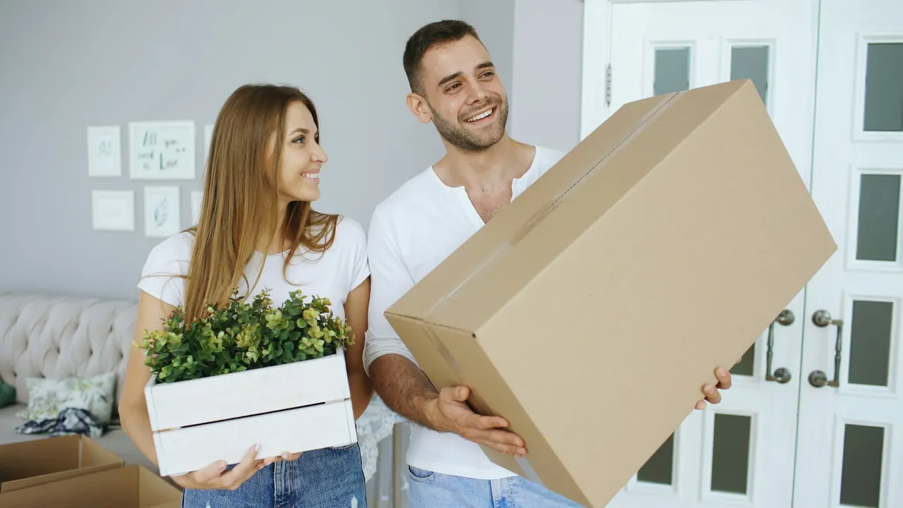 A smiling couple carrying a big moving box and a crate of plants into their new home