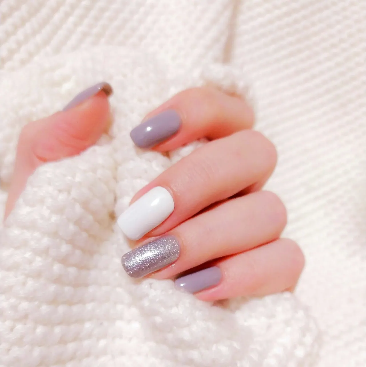A hand with mauve, white and silver glitter nails holding a cosy white knit sweater