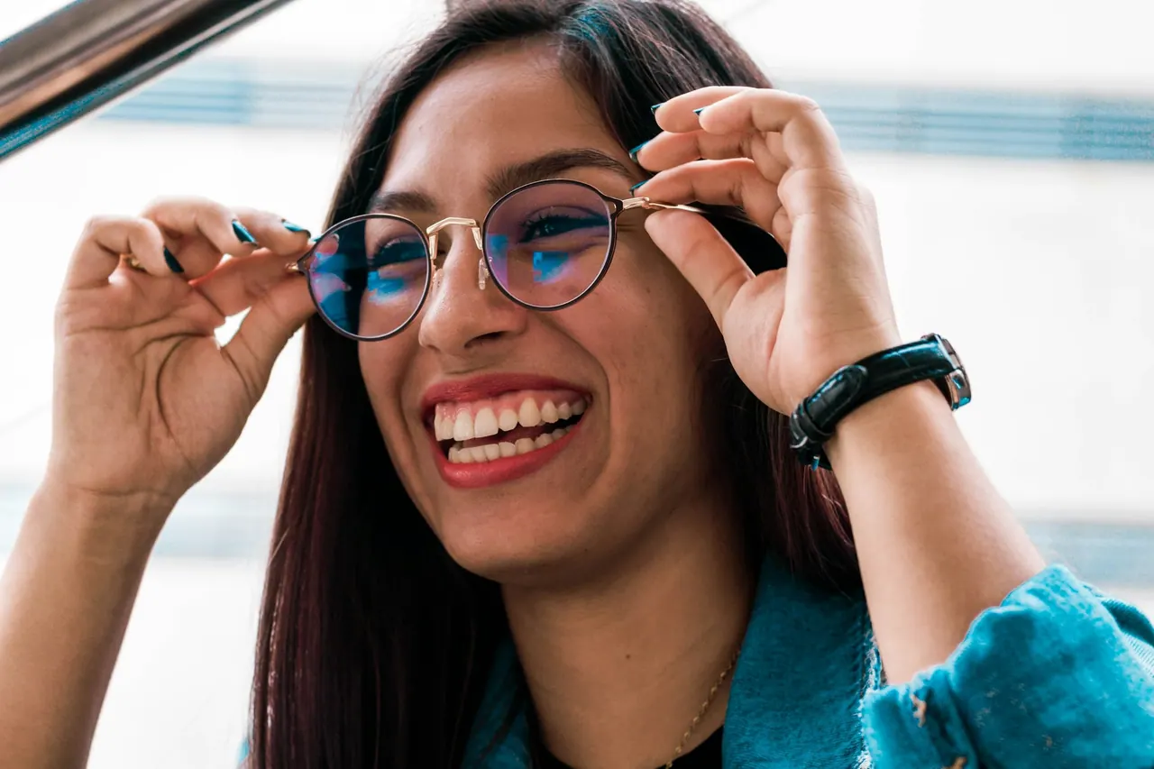 A laughing woman adjusting her round glasses with both hands