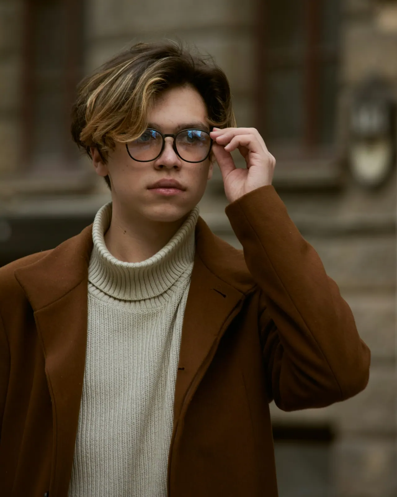A young person in a brown coat adjusting dark framed glasses