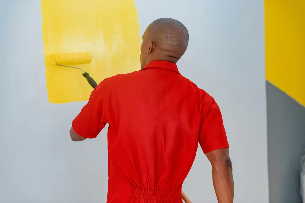 A painter in red overalls rolling bright yellow paint onto a white wall