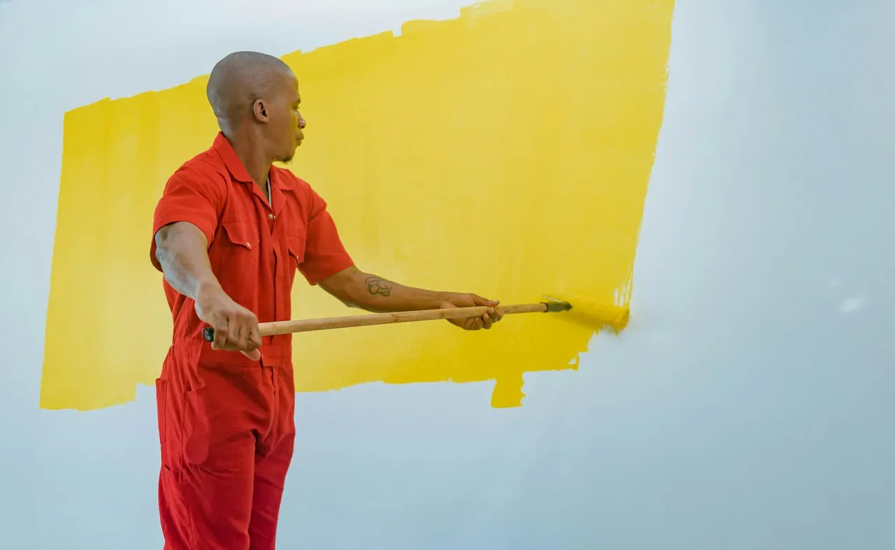 A painter in red overalls rolling bright yellow paint onto a wall