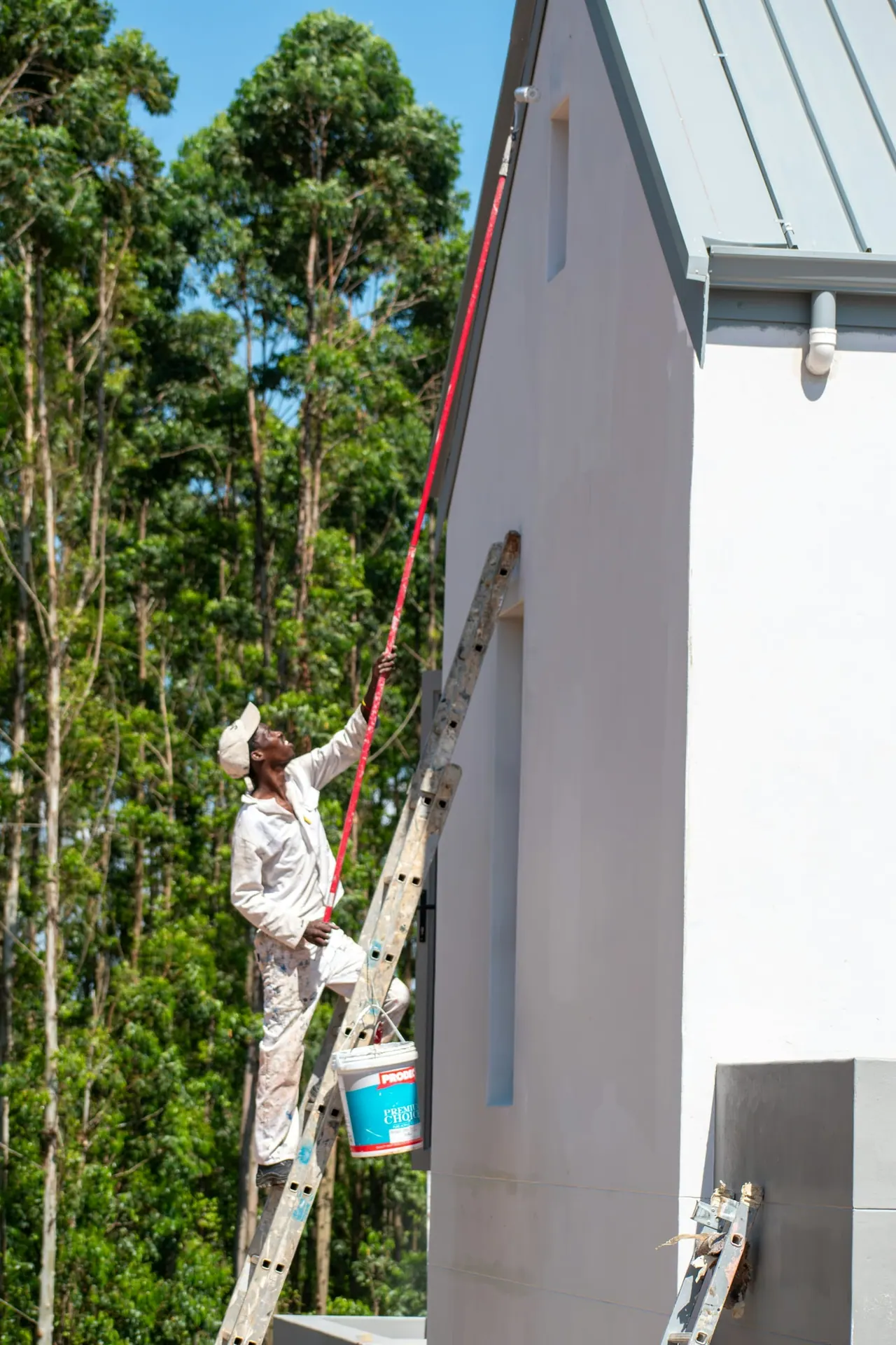 A painter on a tall ladder painting the white gable of a house among trees