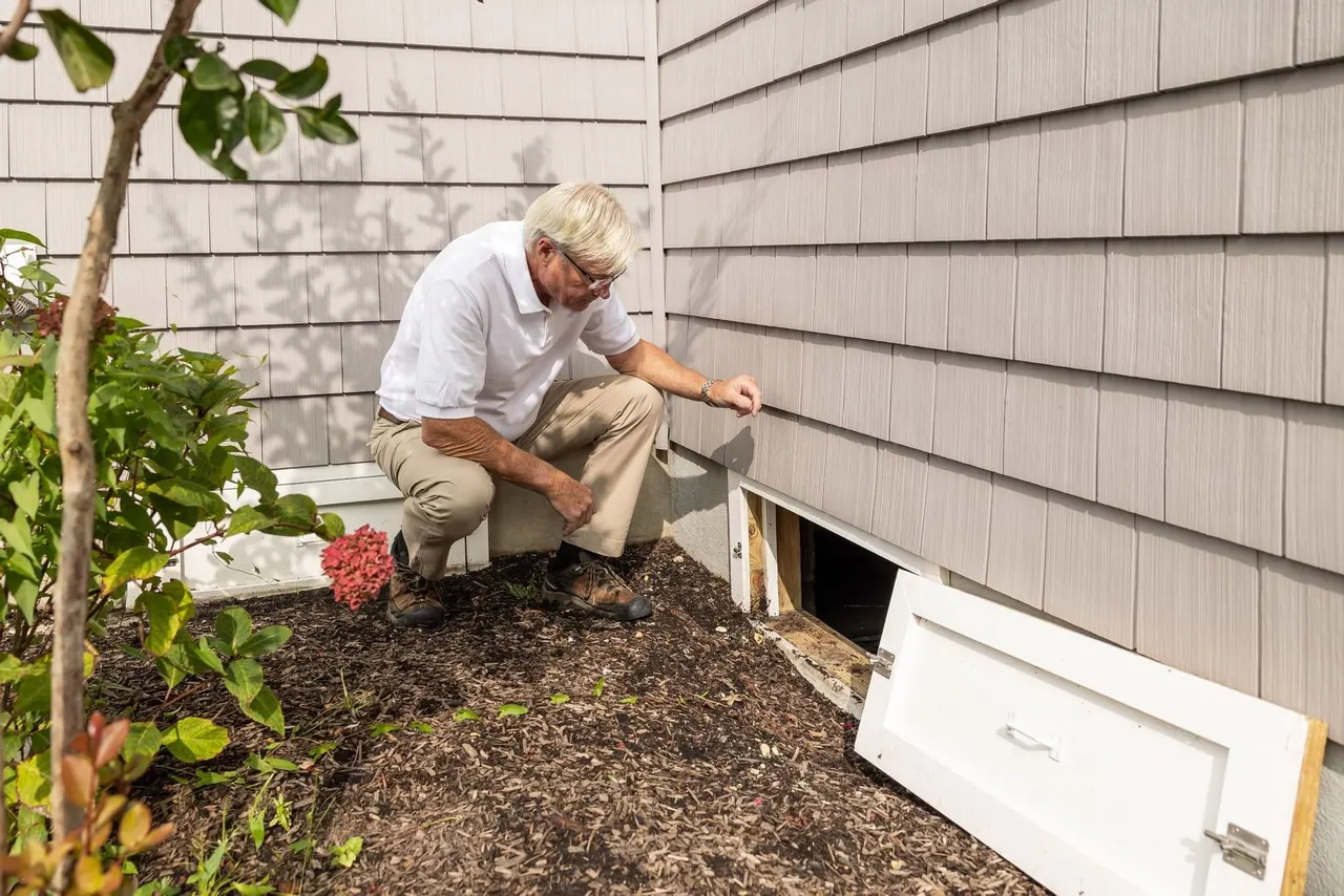 A Brightside technician crouching to inspect the crawl space vent at the base of a house