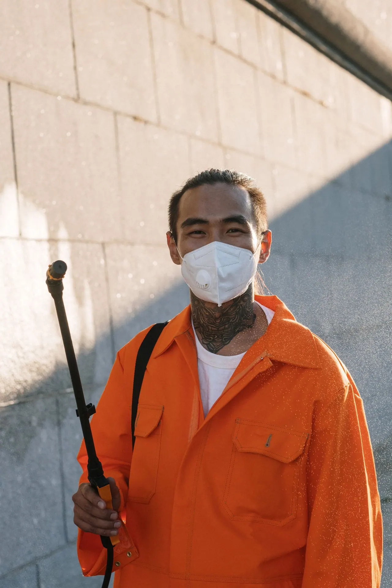 A technician in an orange work jacket holding a sprayer wand, smiling behind a mask