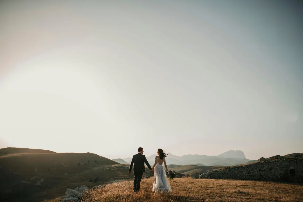 A bride and groom walking hand in hand across a hilltop at dusk