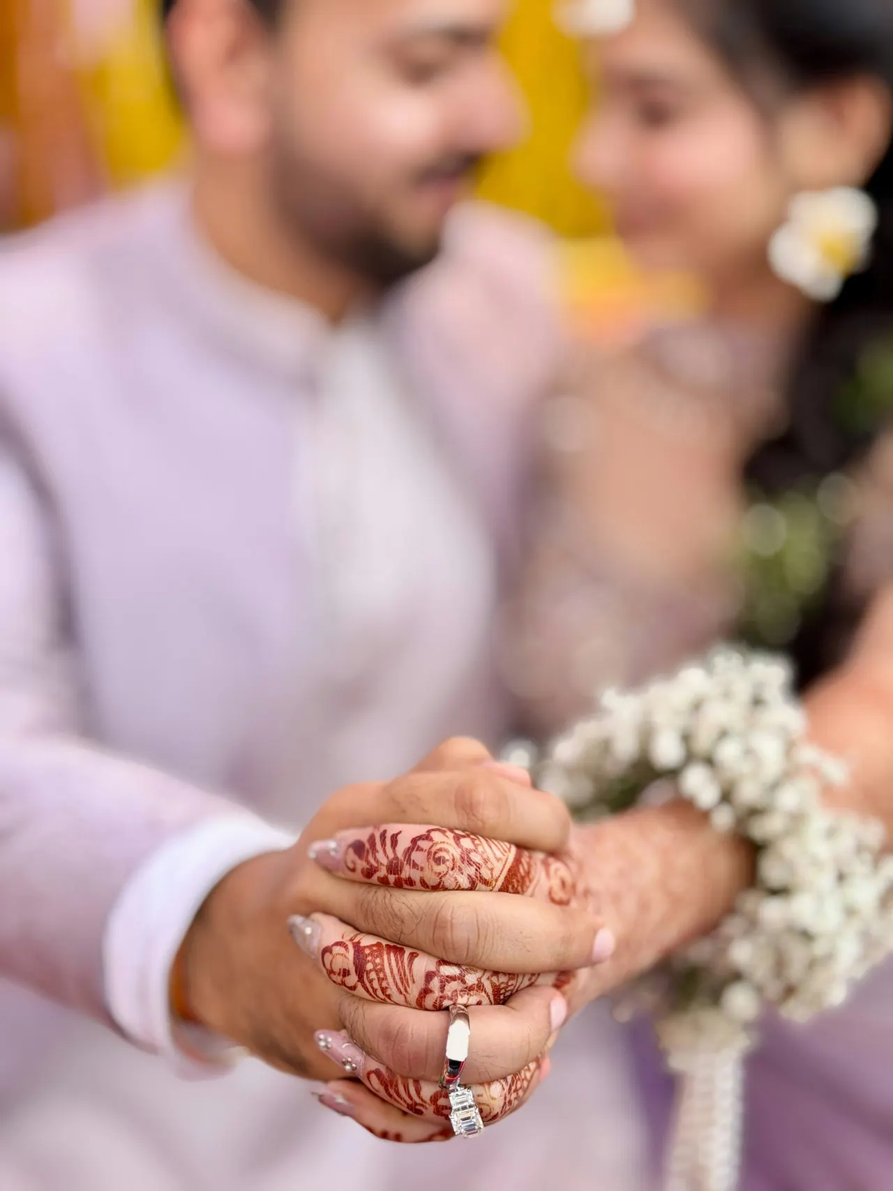 Close up of joined hands with henna patterns and rings, a smiling couple soft behind them