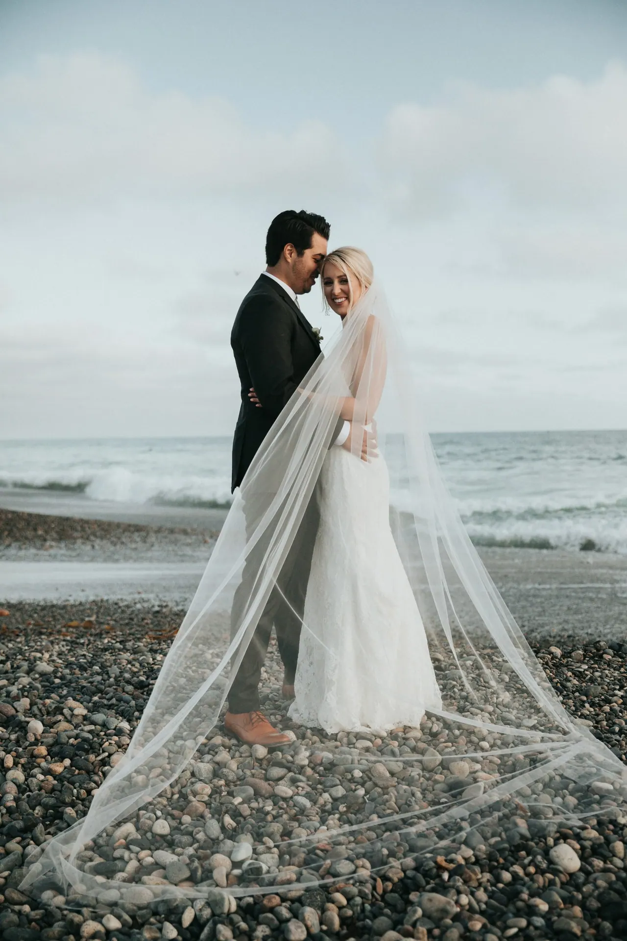 A smiling bride and groom on a pebble beach, her long veil in the wind
