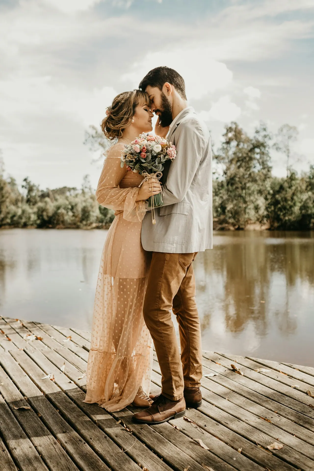 A couple embracing forehead to forehead on a wooden dock by a lake, she holds a bouquet of roses