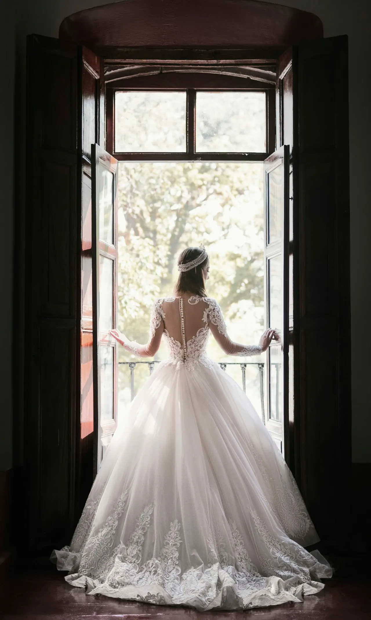 A bride in a lace gown opening tall wooden doors to a sunlit balcony