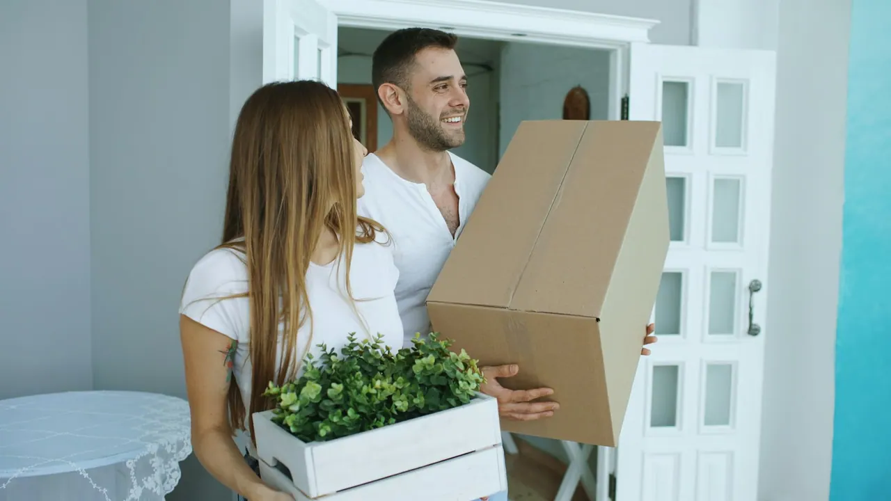 A couple carrying boxes and a plant into their new home