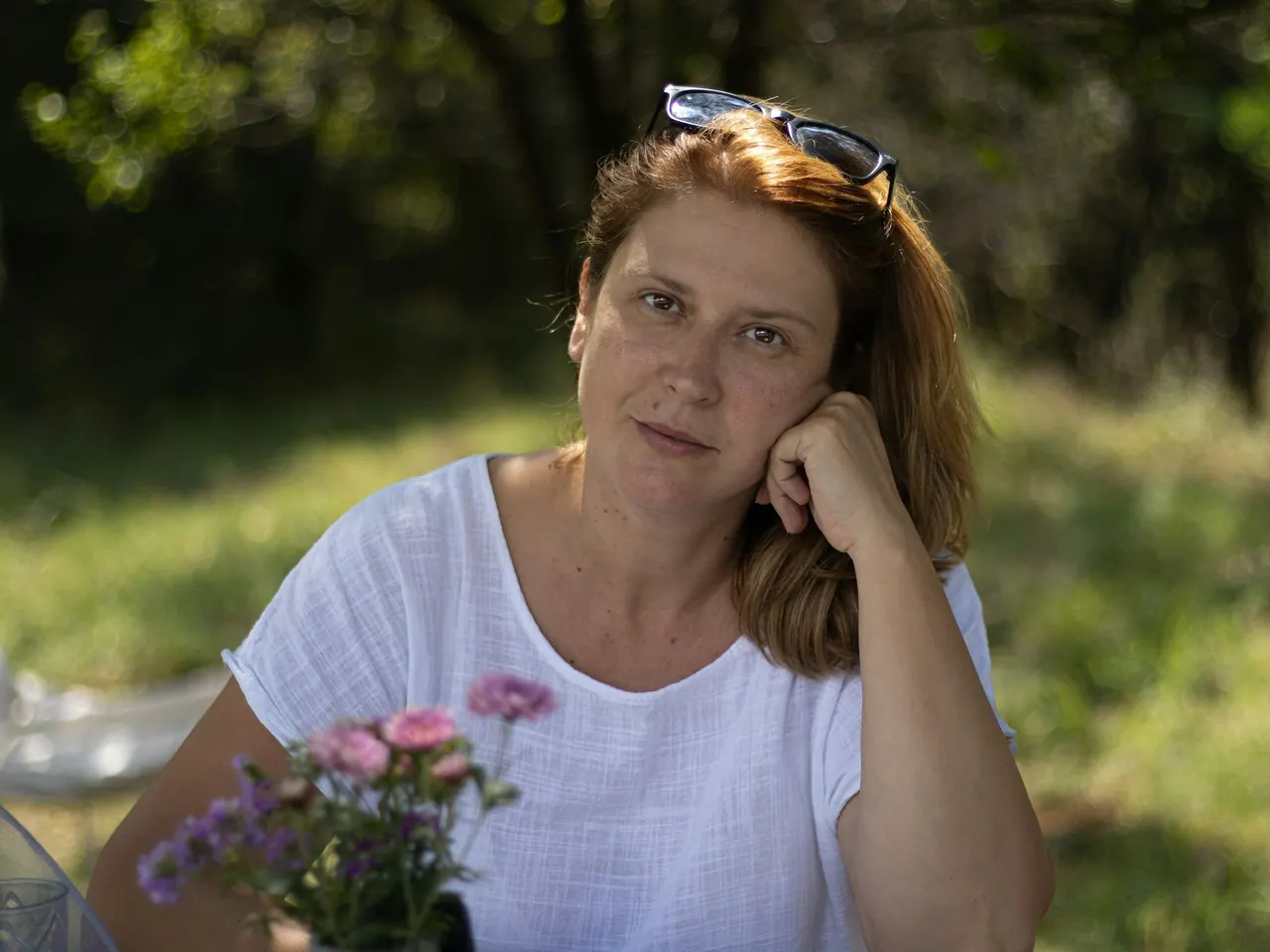 A woman resting her head on her hand at a table outdoors, flowers beside her