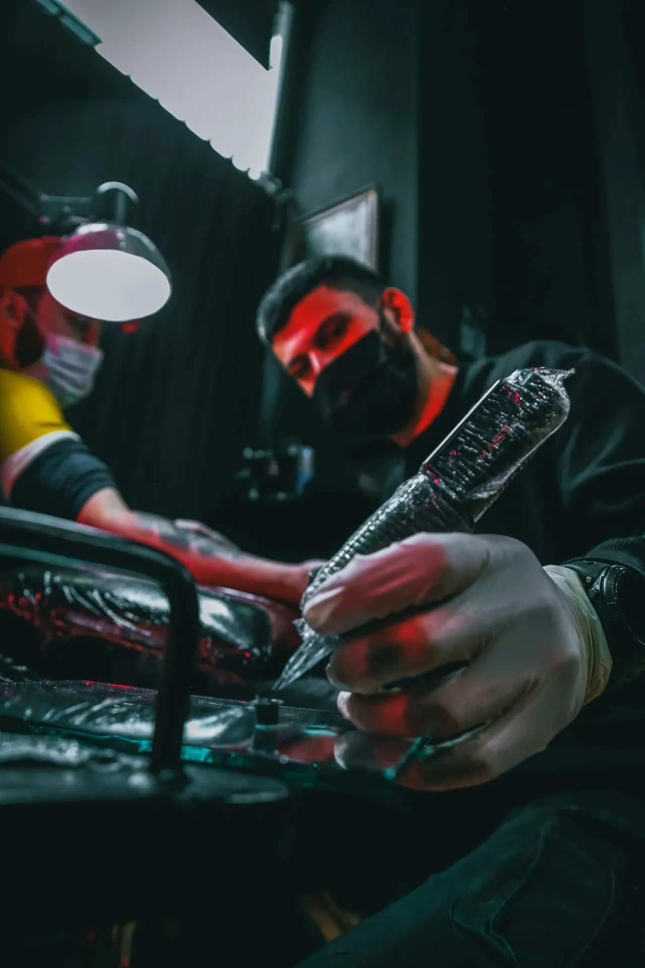 A masked artist tattooing under a lamp in red light