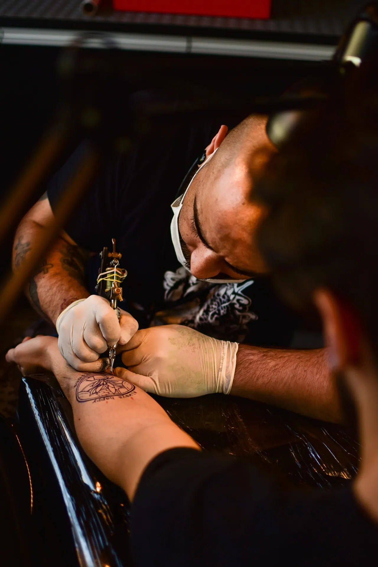 An artist bent over a client, tattooing a forearm