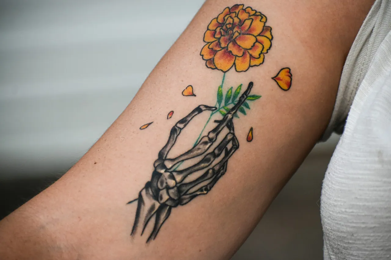 A colour tattoo of a skeleton hand holding an orange marigold