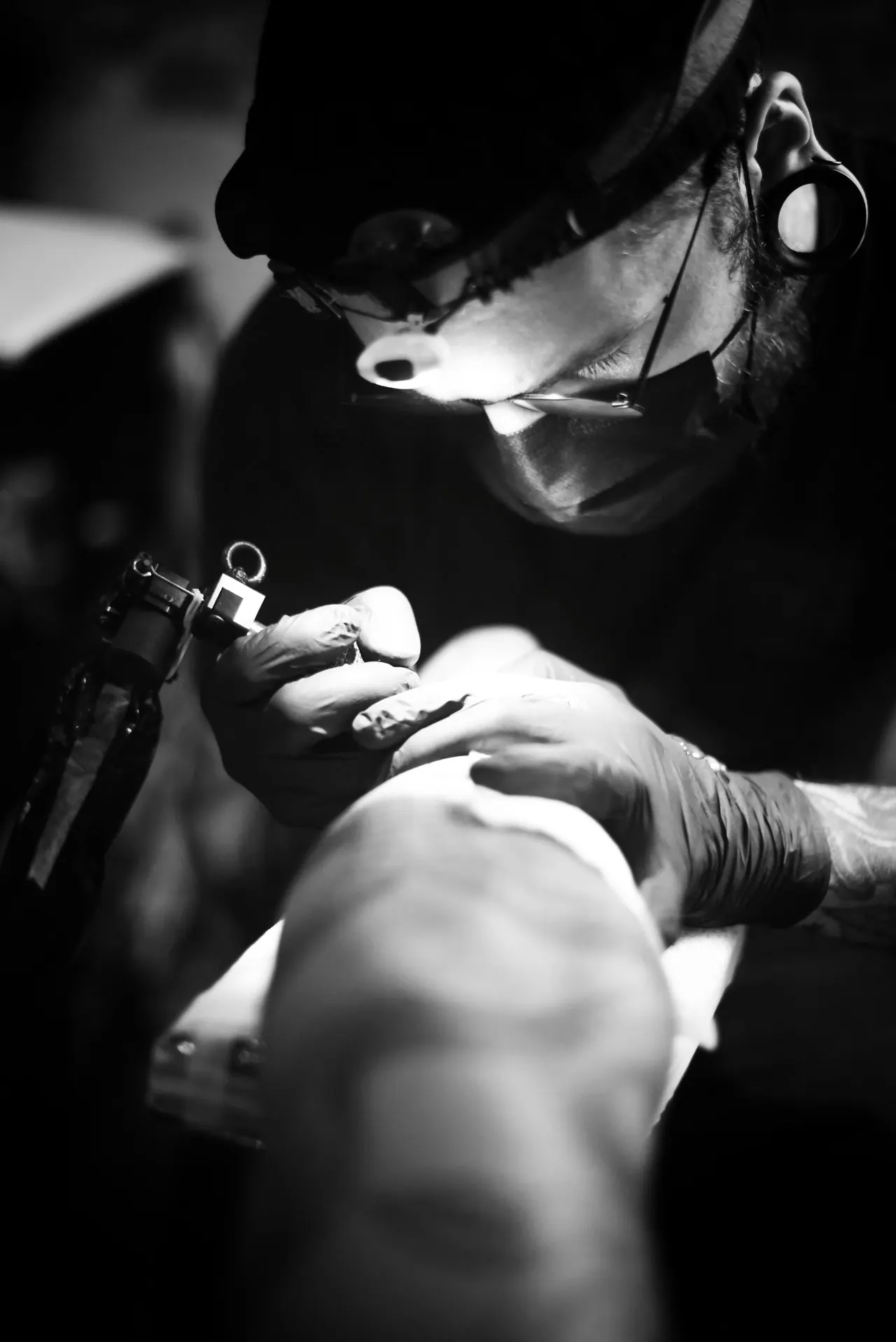 An artist with a head lamp bent close over a tattoo, in black and white