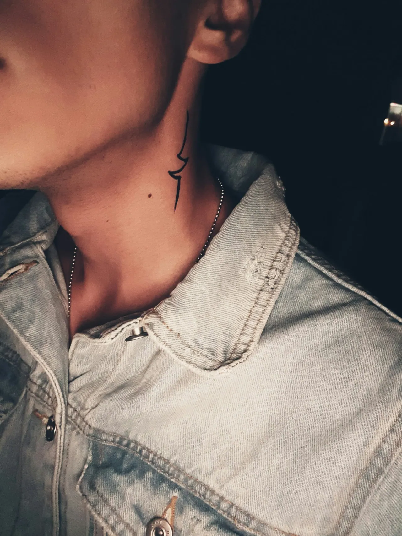 A small fine line tattoo on the side of a neck, above a denim collar